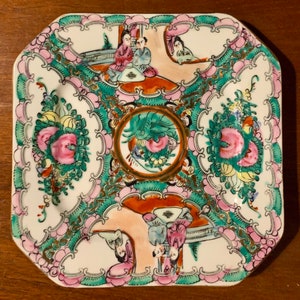 May include: A square porcelain plate with a white background and a colorful floral and figural design. The plate features a central circular motif with a green dragon and pink flowers. The border of the plate is decorated with pink and green floral designs and gold accents. The plate features a scene of people in traditional Chinese clothing.