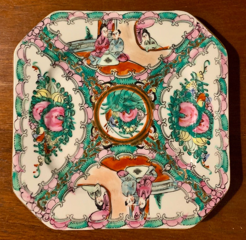 May include: A square porcelain plate with a white background and a colorful floral and figural design. The plate features a central circular motif with a green dragon and pink flowers. The border of the plate is decorated with pink and green floral designs and gold accents. The plate features a scene of people in traditional Chinese clothing.