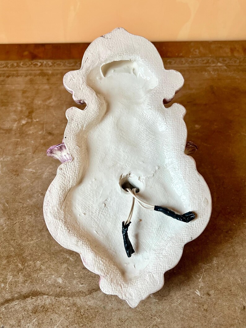 May include: The back of a white ceramic wall sconce with a pink floral design. The sconce has two electrical wires coming out of the back.