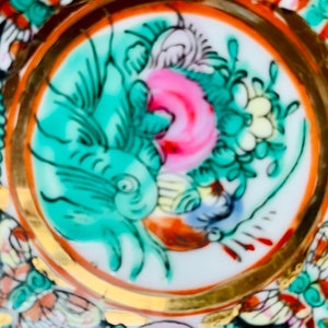 May include: A white porcelain plate with a gold rim and a colorful floral design. The design features a green and blue fish, pink and white flowers, and gold accents.