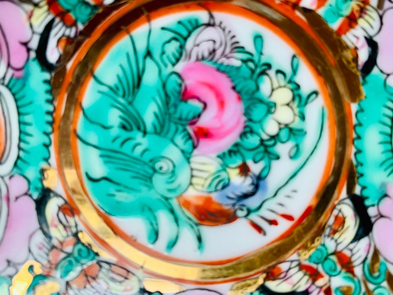 May include: A white porcelain plate with a gold rim and a colorful floral design. The design features a green and blue fish, pink and white flowers, and gold accents.