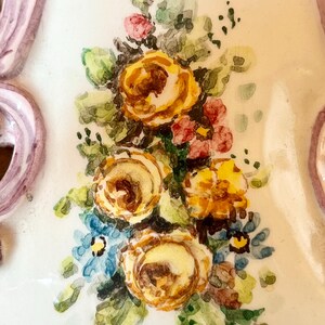 May include: A close-up of a ceramic wall plaque with a painted floral design. The plaque features a bouquet of yellow, orange, and pink roses with green leaves and blue forget-me-nots.