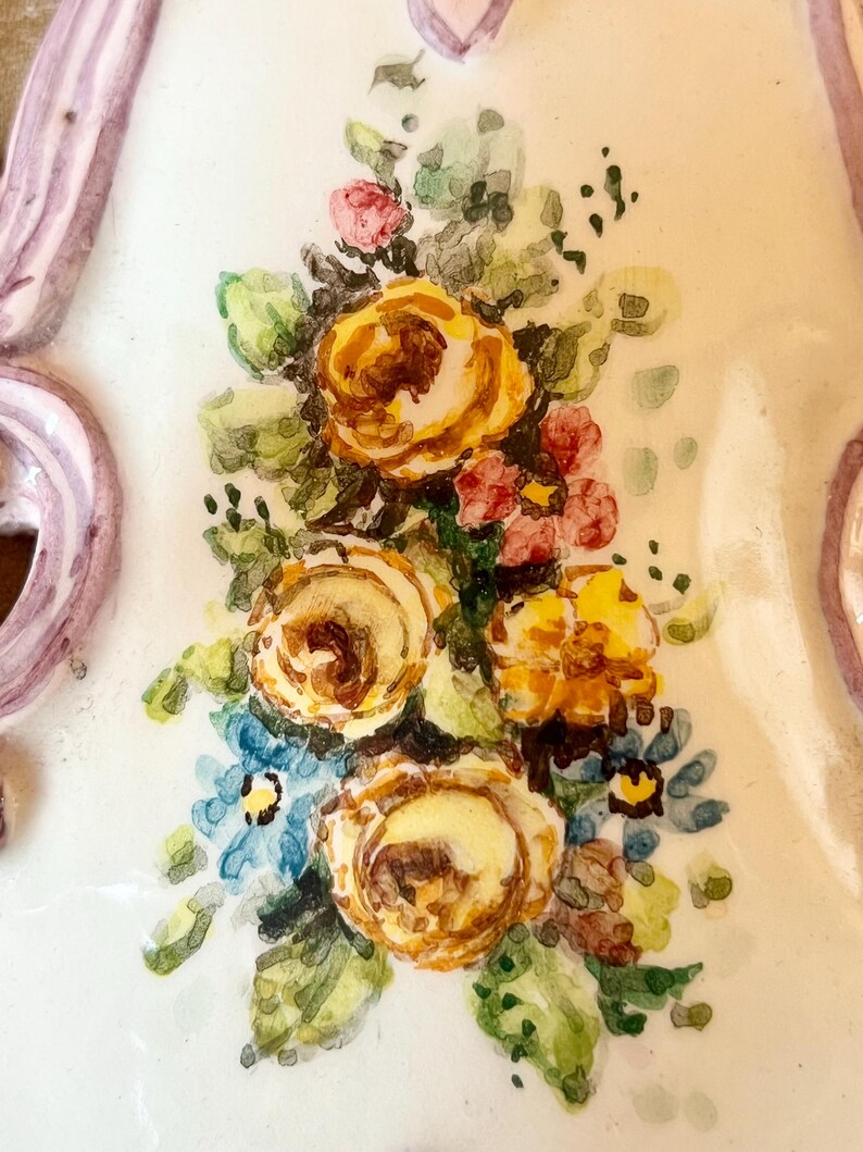 May include: A close-up of a ceramic wall plaque with a painted floral design. The plaque features a bouquet of yellow, orange, and pink roses with green leaves and blue forget-me-nots.