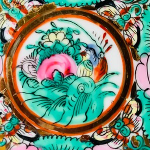 May include: A close-up of a decorative plate with a circular design featuring a green and pink floral motif with gold accents. The plate has a white background and is decorated with a variety of colors, including red, orange, yellow, and blue.