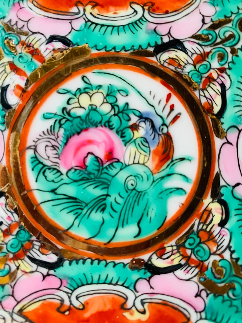 May include: A close-up of a decorative plate with a circular design featuring a green and pink floral motif with gold accents. The plate has a white background and is decorated with a variety of colors, including red, orange, yellow, and blue.