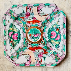 May include: A square porcelain plate with a white background and a decorative design in pink, green, and gold. The design features floral motifs, stylized figures, and intricate patterns.