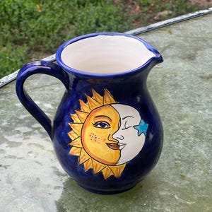 Può includere: Una brocca in ceramica blu scuro con interno bianco. La brocca presenta un disegno dipinto a mano di un sole e una luna sorridenti. Il sole è giallo e arancione, e la luna è bianca con una stella blu. La brocca ha un manico e un beccuccio.
