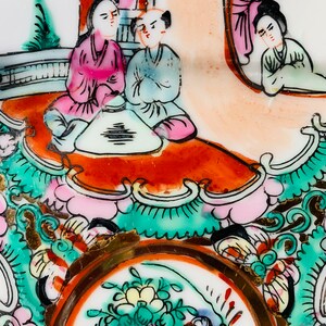 May include: A vintage porcelain plate with a hand-painted design featuring three figures in traditional Chinese clothing. The figures are seated around a table with a floral design. The plate has a gold rim and intricate floral patterns in shades of pink, green, and blue.