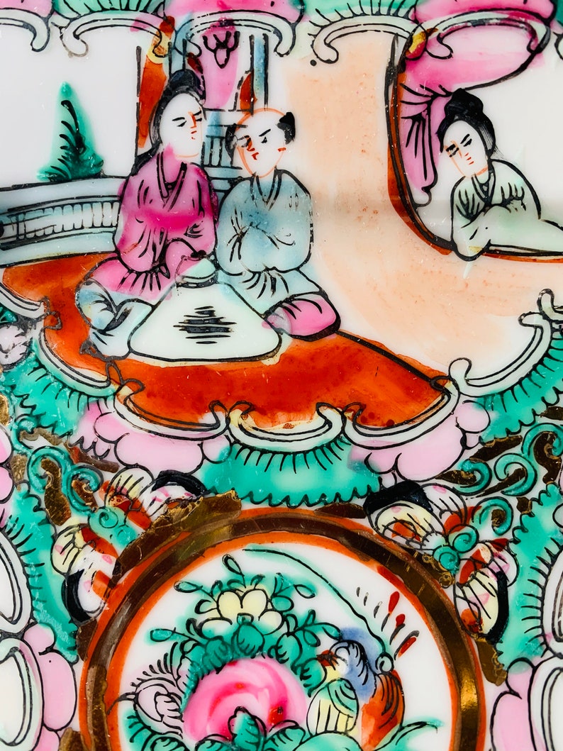 May include: A vintage porcelain plate with a hand-painted design featuring three figures in traditional Chinese clothing. The figures are seated around a table with a floral design. The plate has a gold rim and intricate floral patterns in shades of pink, green, and blue.