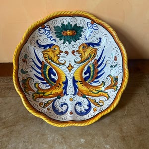 May include: A ceramic plate with a scalloped edge and a yellow rope-like border. The plate features a symmetrical design of two mythical creatures in yellow, blue, and orange, with floral and scroll details on a white background.