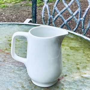 Può includere: Un brocca di ceramica bianca con manico. La brocca è appoggiata su una superficie testurizzata grigia e verde.