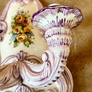 May include: A white ceramic wall sconce with a purple floral design. The sconce has a single candle holder with a scalloped edge.