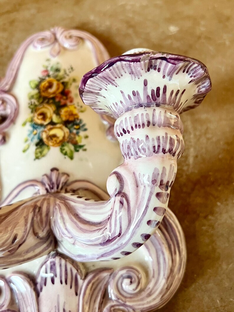 May include: A white ceramic wall sconce with a purple floral design. The sconce has a single candle holder with a scalloped edge.