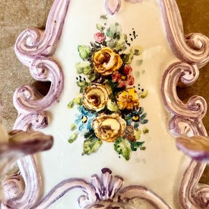 May include: A white ceramic wall sconce with a purple and white floral design. The sconce has a scalloped edge and a floral motif in the center.