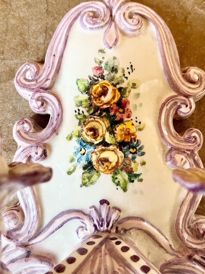 May include: A white ceramic wall sconce with a purple and white floral design. The sconce has a scalloped edge and a floral motif in the center.
