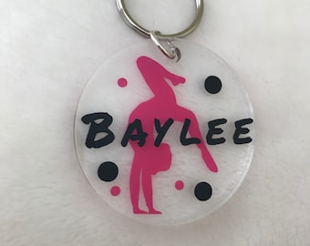 May include: Clear acrylic keychain with a pink gymnast silhouette and the name "BAYLEE" in black lettering. The keychain has a silver key ring and is decorated with black and pink polka dots.