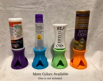 May include: Four 3D printed glue bottle holders in purple, blue, green, and orange. The holders are designed to hold glue bottles upright. The text "More Colors Available" and "Glue is not included" is visible below the holders.
