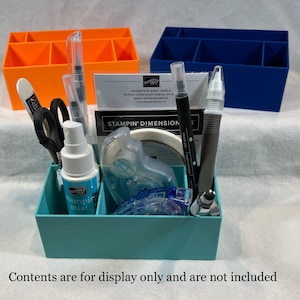 May include: A teal organizer filled with craft supplies, including scissors, spray mist, and pens. Two additional organizers in orange and blue are visible in the background. The text "Contents are for display only and are not included" is at the bottom.