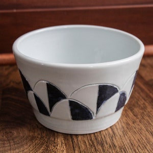 Op de afbeelding: Witte keramische kom met een geometrisch ontwerp in zwart en wit. De kom heeft een afgeronde vorm en een gladde, glanzende afwerking.