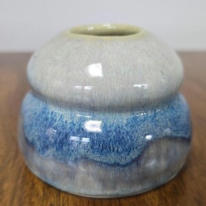 Puede incluir: Un pequeño jarrón de cerámica con un esmalte azul claro y blanco. El jarrón tiene una forma redondeada con una abertura ligeramente acampanada.