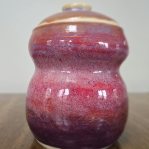 May include: A small, hand-thrown ceramic vase with a rounded, bulbous shape. The vase is glazed in a gradient of pink, red, and purple.