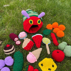 May include: A collection of crocheted items including a large red and green caterpillar head, various fruits like strawberries, oranges, a watermelon slice, a pear, an apple, a pickle, a slice of cheese, a cupcake, an ice cream cone, and a lollipop.