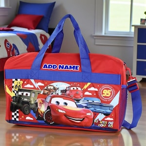 Könnte beinhalten: Rote und blaue Reisetasche mit Lightning McQueen und anderen Charakteren aus dem Film Cars. Die Tasche hat blaue Griffe und einen Schultergurt. Der Text "ADD NAME" ist auf der Tasche aufgedruckt.