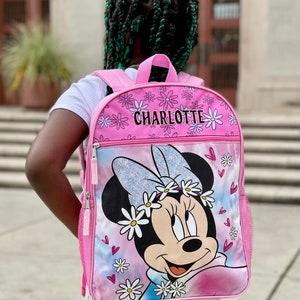 Pu&ograve; includere: Zaino rosa con un design di Minnie Mouse. Lo zaino ha un motivo a margherite bianche e il nome "CHARLOTTE" stampato sul davanti.