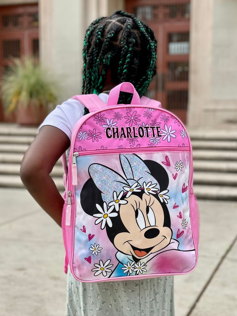 Pu&ograve; includere: Zaino rosa con un design di Minnie Mouse. Lo zaino ha un motivo a margherite bianche e il nome "CHARLOTTE" stampato sul davanti.
