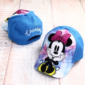 Könnte beinhalten: Blaue Baseballkappe mit einem Batik-Design, das Minnie Mouse zeigt. Die Kappe hat einen weißen Schirm und eine rosafarbene Schleife auf Minnie Mauses Kopf. Auf der Rückseite der Kappe ist der Name "Charlotte" in Weiß gestickt.