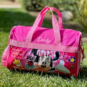 May include: Pink Minnie Mouse themed duffel bag with a zipper closure and a mesh pocket. The bag has a pink strap and the name "Emily" is embroidered on the front.