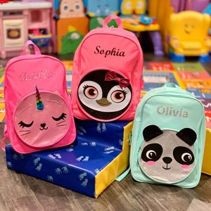 May include: Three pink, turquoise, and teal children's backpacks with animal faces on the front. The pink backpack has a unicorn cat face, the teal backpack has a panda face, and the pink backpack has a penguin face. The backpacks are personalized with the names "Olivia", "Sophia", and "Amelia".