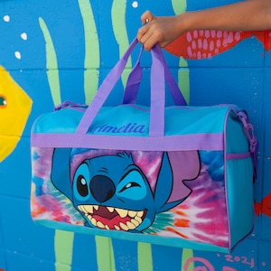 May include: A blue and purple duffel bag with a tie-dye pattern and a cartoon Stitch character. The bag has a purple strap and the name "Amelia" is printed on the front.