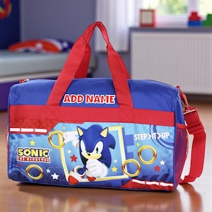 May include: Blue and red Sonic the Hedgehog duffel bag. The bag features a graphic of Sonic, gold rings, and stars. The text "ADD NAME" is above the main graphic. The bag has red handles and a red shoulder strap.