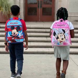 Pu&ograve; includere: Due bambini, uno con uno zaino di Topolino blu e rosso che dice "BENJAMIN" e l'altro con uno zaino di Minnie Mouse rosa che dice "CHARLOTTE", si allontanano da un edificio scolastico.