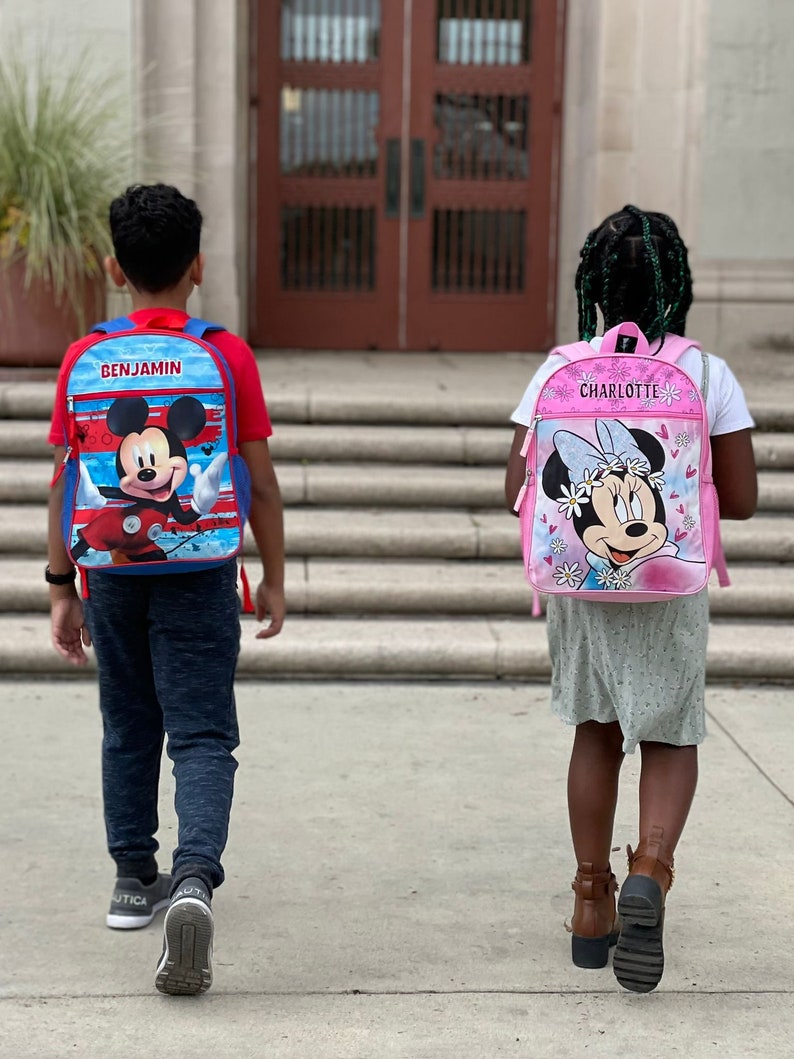 Pu&ograve; includere: Due bambini, uno con uno zaino di Topolino blu e rosso che dice "BENJAMIN" e l'altro con uno zaino di Minnie Mouse rosa che dice "CHARLOTTE", si allontanano da un edificio scolastico.