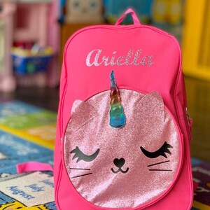 May include: Pink backpack with a unicorn cat face on the front. The backpack has the name "Ariella" embroidered on the front.