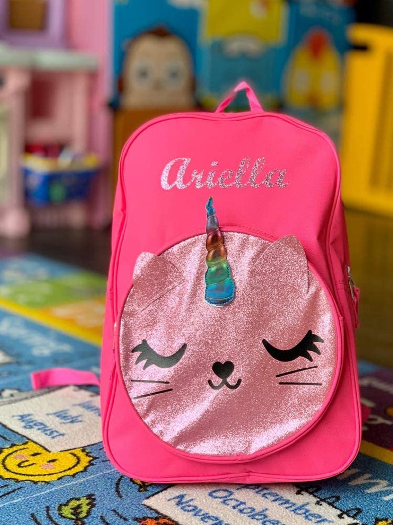 May include: Pink backpack with a unicorn cat face on the front. The backpack has the name "Ariella" embroidered on the front.