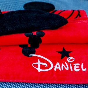 Op de afbeelding: Rode strandhanddoek met een zwart Mickey Mouse-ontwerp en de naam "Daniel" in witte letters.