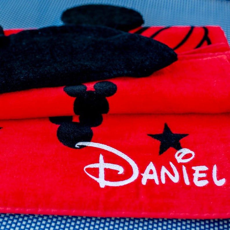 Op de afbeelding: Rode strandhanddoek met een zwart Mickey Mouse-ontwerp en de naam "Daniel" in witte letters.