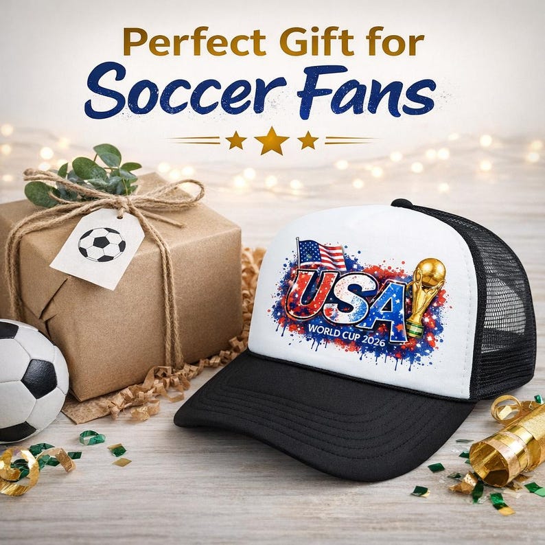 Puede incluir: Gorra de camionero en blanco y negro con un panel frontal blanco con un dise&ntilde;o de la Copa del Mundo de EE. UU. 2026. El dise&ntilde;o incluye la bandera estadounidense, la palabra "USA" y un trofeo dorado. Tambi&eacute;n hay un bal&oacute;n de f&uacute;tbol y una caja de regalo en la imagen.