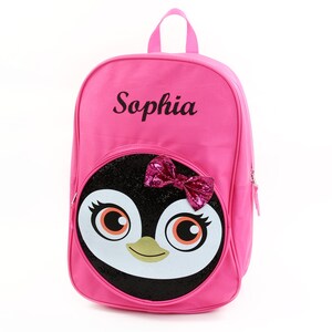 May include: Pink backpack with a penguin face on the front. The penguin has a sparkly black body, white face, and a pink bow. The backpack has the name "Sophia" embroidered on the front.