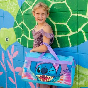 May include: A blue and purple duffel bag with a tie-dye pattern and a cartoon Stitch character. The bag has the name "Amelia" written on it.