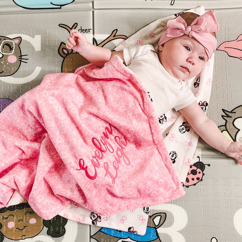 K&ouml;nnte beinhalten: Eine rosa Babydecke mit wei&szlig;en Punkten und dem Namen "Evelyn Leigh", der in Rot gestickt ist. Die Decke ist gefaltet und liegt auf einer grau-wei&szlig;en Spielmatte.