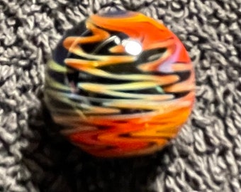 May include: A spherical glass marble featuring a swirling pattern of black, bright orange, and pale yellow colours. The surface is smooth and reflective, with light catching the curved glass to create a bright white highlight on the top.