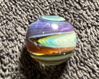 May include: A colorful glass marble with swirling patterns of purple, blue, yellow, and brown. The marble is round and has a glossy finish, reflecting light. This decorative marble is a unique collectible.