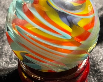 May include: A colorful glass marble with swirling patterns of red, yellow, blue, and turquoise. The marble is displayed on a dark wooden stand, creating a striking contrast. The marble's design is reminiscent of a galaxy.