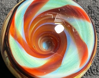 Puede incluir: Una canica de vidrio redonda y colorida con un patrón en espiral. El diseño presenta bandas de color naranja, turquesa y azul claro, creando un efecto de espiral. La canica tiene un acabado brillante y un vórtice central.