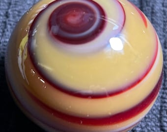 May include: A glass marble with a spiral design. The marble has a yellow base with swirling red and white bands. The centre of the marble has a deep red spiral. The marble is round and has a glossy finish.