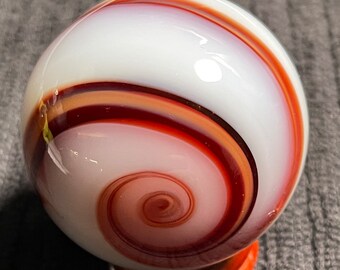 May include: A close-up of a glass marble with a white base and swirling patterns of red, orange, and brown. The marble has a spiral design in the center and is resting on an orange stand. The marble is a collectible item.